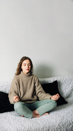Young woman sitting in lotus position on the sofa with her hands on her knees, meditating, trying to relax her mind and relieve stressの写真素材