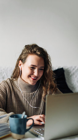 Student video chatting with friends, taking a break from studying at home. Young woman in headphones sitting at kitchen table with laptop and coffee, smiling while chatting with friends on video callの写真素材