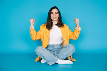 Vibrant Portrait of a Joyful Girl Celebrating Success with Raised Fists and a Victorious Yes  - Isolated on Blue Background - Positive, Optimistic Emotionsの写真素材
