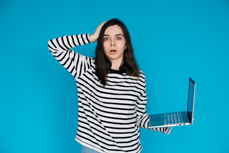 Emotional Young Woman in Striped Sweater Distraught by Bad News on Laptop - Expressive Female Feeling Sad and Disheartened - Isolated Blue Background - Perfect for Conceptual, Emotionalの写真素材