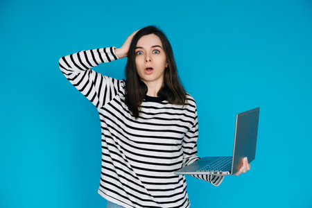 Emotional Woman in Striped Sweater Reacting to Bad News on Laptop - Conceptual Image of Sadness, Disappointment, and Technology - Isolated Blue Background with Copy Space - Perfect for Emotionalの写真素材