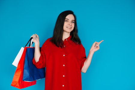 Cute Woman in Red Shirt with Shopping Bag - Pointing to Empty Space - Perfect for Promoting Sales and Discounts - Isolated on Blue Backgroundの写真素材