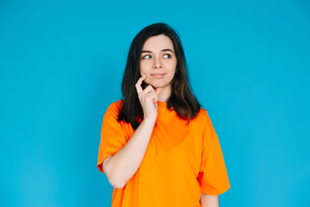 Attractive Young Woman in Trendy Orange Clothes, Pensive and Skeptical Expression, Empty Space for Copy, Isolated on Blue Background - Portrait Photoの写真素材