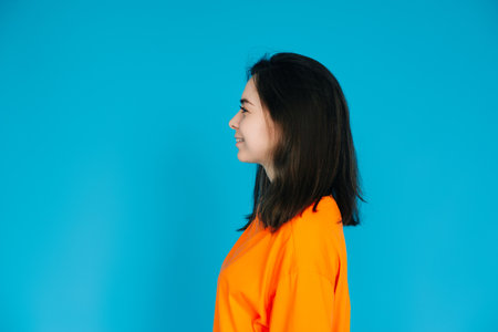 Radiant Young Woman in Stylish Orange Attire Smiling with a Beaming Toothed Smile, Profile Portrait, Isolated on Blue Backgroundの写真素材