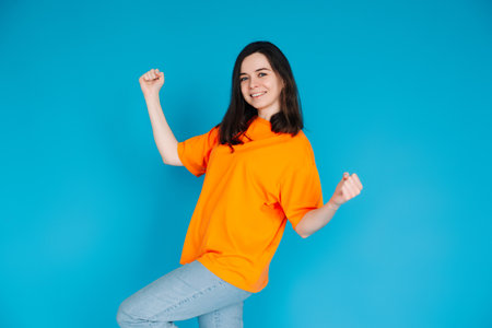Excited Young Woman Celebrating Huge Sales Promotion, Raising Fists in Victory, Trendy Orange Outfit, Isolated on Blue Background, Portrait Photoの写真素材