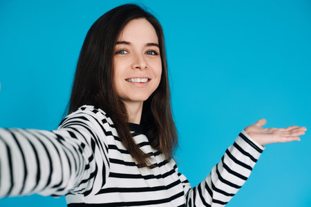 Cheerful Girl Taking Selfie with Empty Space, Adorable Young Woman with Toothsome Smile, Demonstrating with Arm and Palm, Isolated on Blue Background, Portrait Photoの写真素材