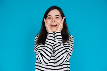Expressive Portrait of a Surprised and Joyful Young Woman with Palms on Cheekbones, Isolated on Blue Backgroundの写真素材
