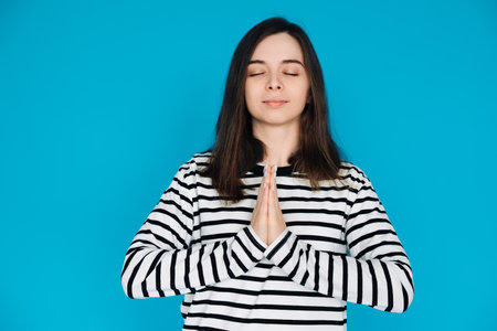 Serene Portrait of a Beautiful Young Woman Meditating with Closed Eyes and Palms, Inner Peace and Relaxation, Isolated on Blue Backgroundの写真素材