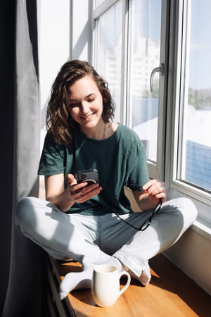 Lockdown Lifestyle: Young Woman Using Smartphone at Home, Engaging in Relaxation and Connection While Looking Out of Window - Technology in Leisure Timeの写真素材