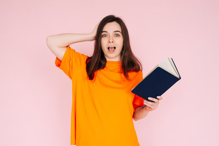 Captivating Reading Experience: Surprised Woman with Hand on Head, Engrossed in a Book with an Intriguing Genre - Fascination and Immersion Concept on Pink Background.の写真素材