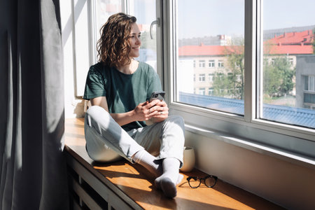 Digital Disconnect: Peaceful Young Woman Finds Serenity at Home, Gazing Out of Window, Balancing Technology and Relaxation in a Lockdown Eraの写真素材