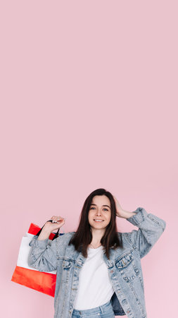 Excited young woman in casual denim shirt raises her arm in celebration, standing in front of a pink background, happy after shopping or finding a great deal online. Vertical photographyの写真素材