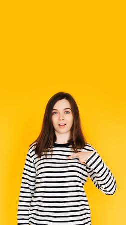 Delighted Woman Pointing at Herself in Amazement - Humorous Reaction Concept on Bright Yellow Background. Surprised and Amused Woman Pointing at Herself with Open Mouth - Self-Recognition Humorの写真素材