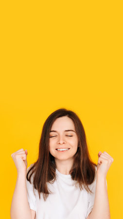 Exhilarated Woman Celebrating Big Win with Raised Fists - Excitement, Victory, and Financial Success Concept on Vibrant Yellow Backgroundの写真素材