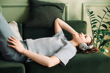 A smiling middle-aged woman wearing wireless headphones listens to music while lying on the sofa. Blissful Melodies. Middle-Aged Woman Enjoys Music with Wireless Headphones.の写真素材