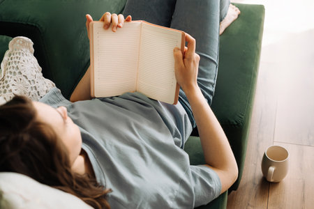 Leisure Time. Young Woman Enjoying Book, Meal, and Tea on Living Room Sofa. Relaxed Reading and Dining in Home Setting: Cozy Lifestyle with Books, Food, and Beverage. Relaxation in Domestic Comfort.の写真素材