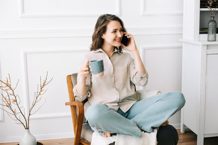 Cheerful Young Woman Talking on Smartphone, Relaxing in Armchair with Drink. Happy Young Woman Talking on Smartphone, Relaxing with Drink in Armchair.の写真素材