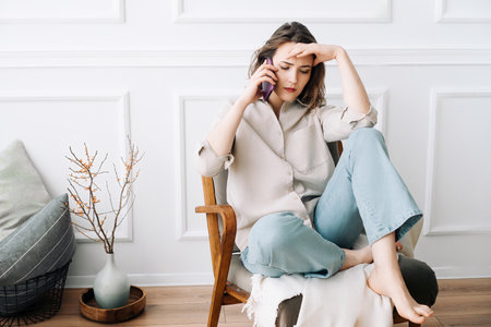 Upset Millennial Woman on Phone, Conveying Grief, Seated on Chair, Reacting to Unsettling News. Despaired sad millennial female calls by phone, talks, sits on chair, suffering from bad news.の写真素材