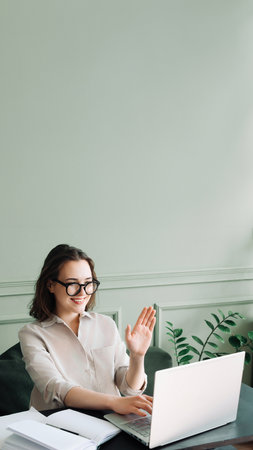 Happy Young Woman in Glasses Engaged in Joyful Laptop Video Chat. Energetic Video Chat: Joyful Woman in Glasses Engages in a Laptop Call, Waving Cheerfully.の写真素材