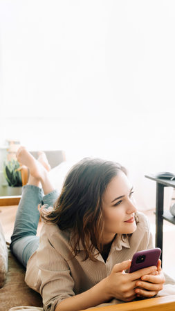 Contemporary Bliss. Joyful Millennial Woman on Sofa, Embracing Leisure, Engrossed in Phone, Gazing into Tranquil Living Room Space. Relaxation, Technology, and Comfortable Living Concept.の写真素材