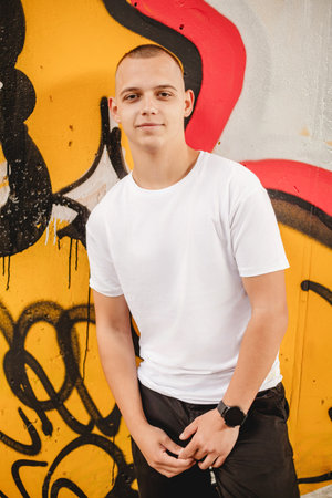A young man stands in front of a graffiti wall, wearing a white shirt and black shorts. He is...