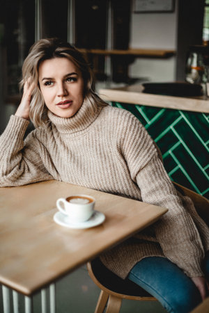 Thoughtful Woman with Coffee in Cozy Cafe. A woman in a sweater sits at a cafe table, hand on her...