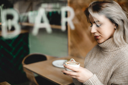 Woman sipping coffee at a cafe table, surrounded by a warm, inviting atmosphere. Ideal for images of casual cafe life, relaxation, and enjoying coffee in a comfortable setting.の写真素材