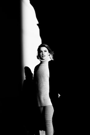 Woman wearing a white sweater and black pants stands against a wall, smiling warmly at the camera....