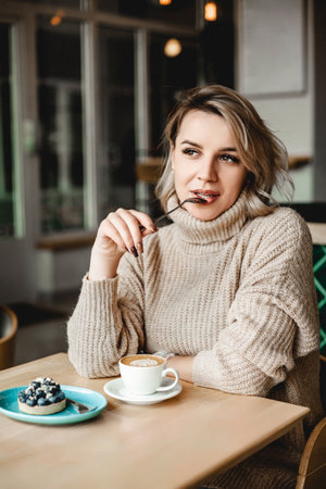 A woman is sitting at a table with a cup of coffee and a pastry. She is wearing a sweater and has her hands in her mouth. The scene is casual and relaxed, with the woman enjoying her coffeeの写真素材