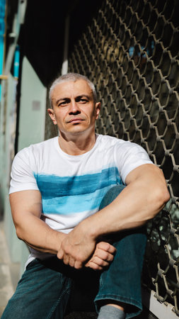 A man with a blue and white striped shirt sits on the ground. He has a tattoo on his arm and a...