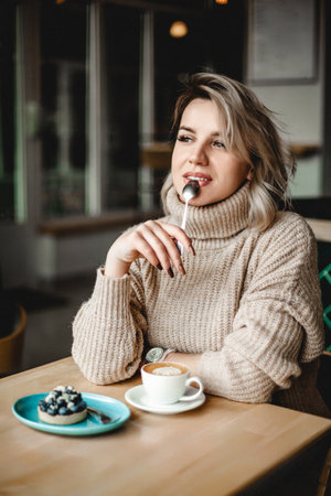 A woman is sitting at a table with a cup of coffee and a pastry. She is wearing a sweater and has a spoon in her mouthの写真素材
