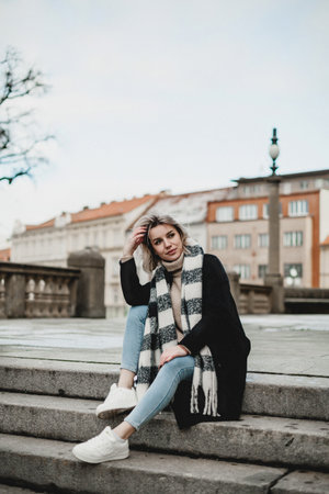 A woman is sitting on a set of stairs wearing a black coat and a white scarf. She is smiling and looking at the cameraの写真素材