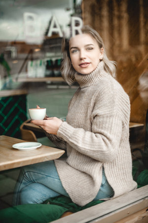 A woman is sitting at a table with a cup of coffee in front of her. She is wearing a sweater and jeansの写真素材