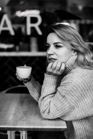 A woman sits at a table with a cup of coffee in front of her. She is wearing a sweater and has her hands on her lap. The scene is set in a cafeの写真素材
