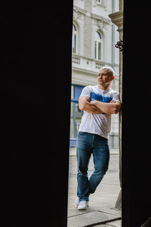 A man in a white shirt and blue jeans stands in front of a building with his arms crossedの写真素材