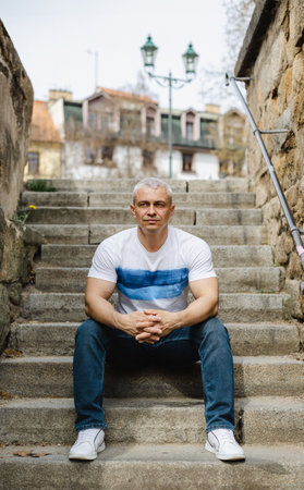 A man in a white shirt and blue jeans sits on a set of stairs. He is looking at the camera with a serious expression. Concept of solitude and contemplationの写真素材