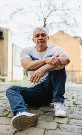 A man with tattoos on his arm sits on the ground with his hands on his knees. He is wearing a white shirt and blue jeansの写真素材