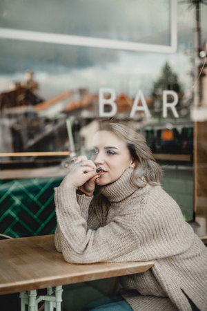 A woman is sitting at a table in a bar, looking out the window. She is wearing a sweater and has her hands on her faceの写真素材