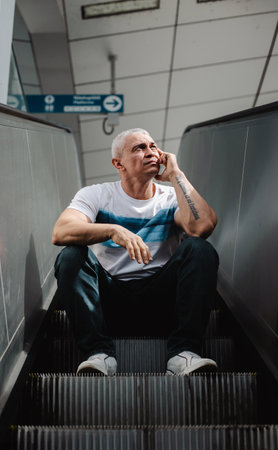 A man is sitting on a set of stairs and talking on his cell phone. He is wearing a white shirt and blue jeansの写真素材