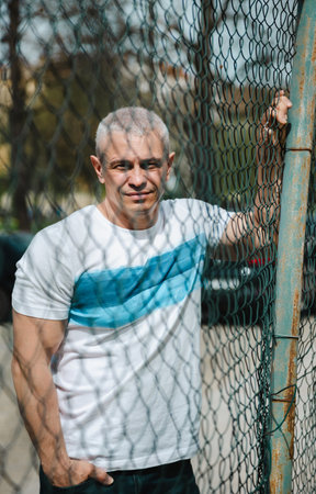 A man stands in front of a chain link fence. He is wearing a white shirt with blue stripes