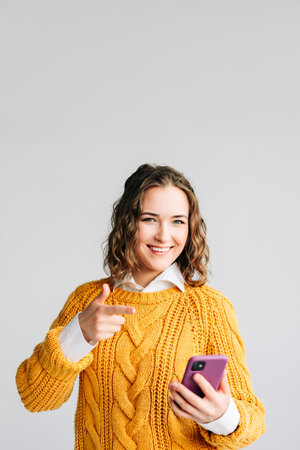 A smiling young woman model points at her phone against a white background. She showcases a new app, advertising mobile web services, and promoting shopping promotions with enthusiasm.の写真素材