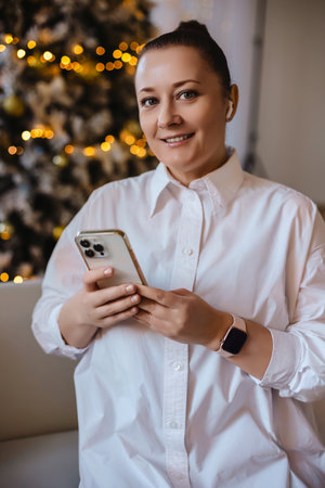 A cheerful woman in a white shirt holds her smartphone while sitting by a beautifully lit Christmas tree. Perfect for holiday lifestyle, technology, or festive content themes.の写真素材