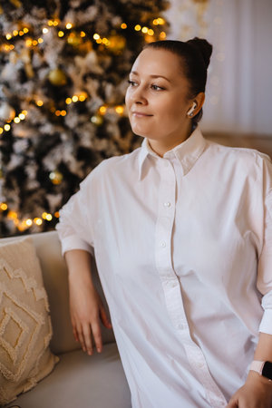 A serene woman in a white shirt rests on a couch with a decorated Christmas tree in the background. Warm lighting and cozy details create a peaceful holiday atmosphereの写真素材