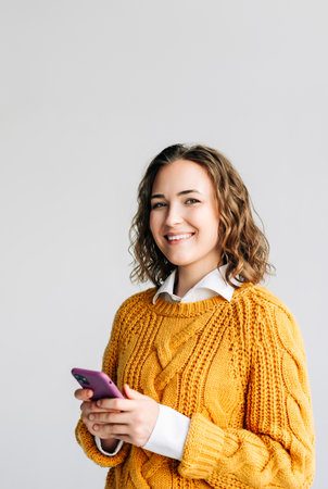 Smiling young woman with curly hair using her smartphone. She is the browsing internet, standing against a white background. Perfect for illustrating modern communication and technology usage.の写真素材