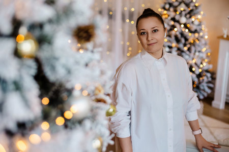 A calm woman stands near a decorated Christmas tree with golden ornaments and festive lights. The soft bokeh effect enhances the cozy holiday atmosphere of the interior.の写真素材