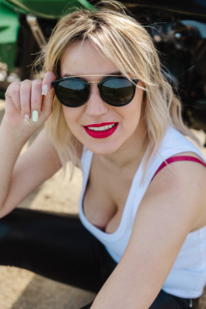 A young woman with blonde hair wearing stylish sunglasses and red lipstick smiles while sitting on the ground. Her confident look and relaxed pose make the photo ideal for fashion and beauty themes.の写真素材