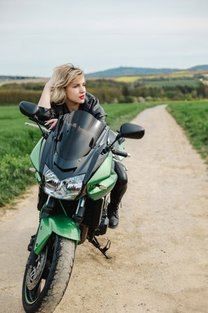 A stylish woman with red lipstick sits on a green motorcycle parked on a dirt road, wearing a black leather jacket. She gazes at the scenic view, evoking freedom and adventure.の写真素材