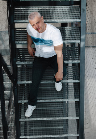 A man in a white shirt and blue jeans is sitting on a set of stairs. The stairs are made of metal and are quite tall. The man is posing for a photo, and the overall mood of the image is casualの写真素材