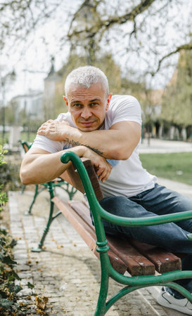 A man is sitting on a green bench in a park. He is wearing a white shirt and blue jeansの写真素材