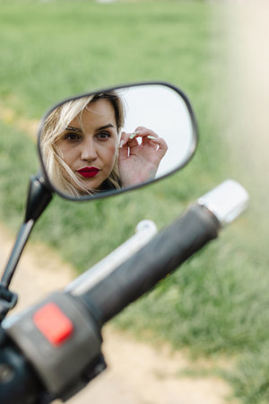 Woman with Red Lipstick Looking into Motorcycle Mirror. A woman with red lipstick gazes into a black and silver motorcycle mirror. Perfect for themes of reflection, biker style, and modern fashionの写真素材